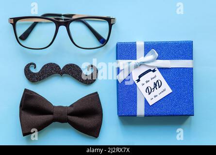Close-up of glasses, mustache, tie and gift with the inscription happy father's day to the best dad in the world on a blue background.  Stock Photo