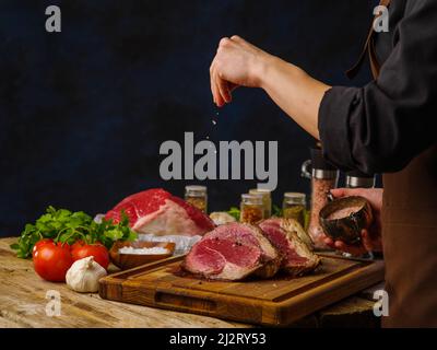 Steaks from beef, pork, cooked by the hands of a professional chef on a wooden cutting board. The chef is salting the steaks. Salt in frozen flight. V Stock Photo