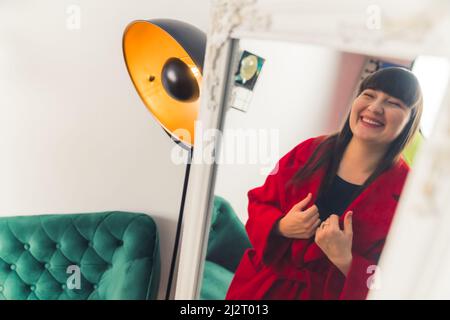 Young European woman trying on red coat. looking at the mirror and smiling online shopping concept portrait copy space . High quality photo Stock Photo