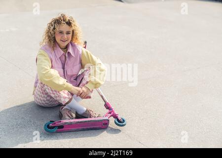 Attractive young fresh-faced blond hipster woman sits on the sports ground smiling, legs crossed, pink scooter in front of her look at the camera full shot copy space. High quality photo Stock Photo