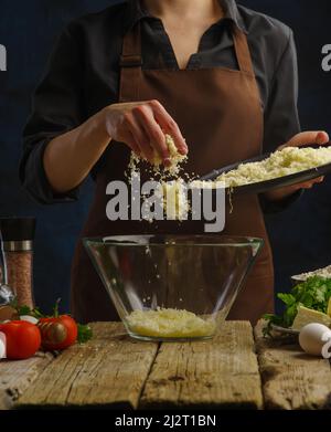 The chef prepares dishes from grated cheese. He tosses the cheese into a large glass bowl. Vegetables, herbs, spices lie on a wooden table. Recipes fo Stock Photo