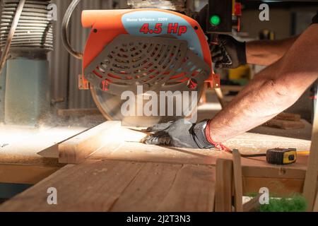 hands of a young carpenter working on a circular saw machine cutting a wooden board in a carpentry workshop using safety black gloves Stock Photo
