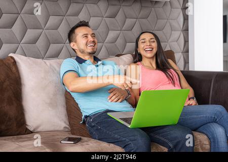 Latin man and woman in their 30s laughing fun looking at content on their laptop sitting in the living room of their home Stock Photo