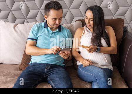 Young couple of man and pregnant woman making purchases online on their cell phone with their credit card sitting in the living room of the house Stock Photo