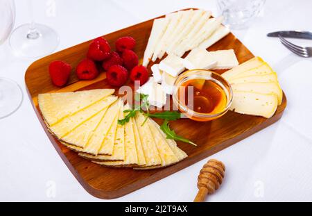 Slices of artisanal cheeses on wooden board with raspberries and honey Stock Photo