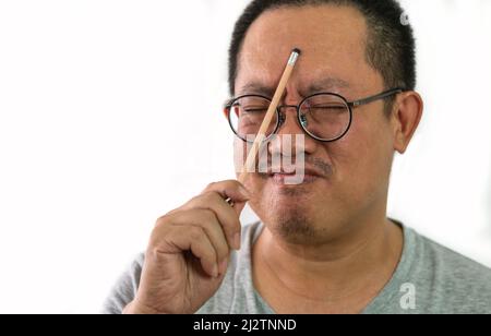 Asian middle-aged man with gray tee shirt having expression of hard thinking on his face, holding a pencil touching forehead, wearing eyeglasses, clos Stock Photo