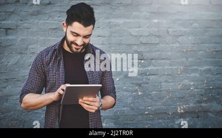 If its new hell know about it. Shot of a young man standing outdoors and using a digital tablet against a gray wall. Stock Photo