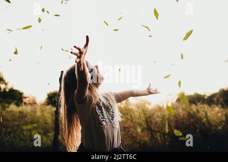 Happy little blonde girl plays with green leaves in garden, field. The child smiles, laughing and has fun in park. Playful kid throwing up leaves in summer or autumn season. Child lays in country side Stock Photo