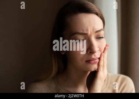 Toothache and dentistry. A beautiful young woman suffers from terrible severe pain in her teeth, touching her cheek with her hand. Dental care and hea Stock Photo