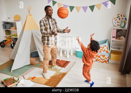 Full length portrait of young black father playing with toddler son indoors and throwing basketball ball in cozy kids room interior Stock Photo