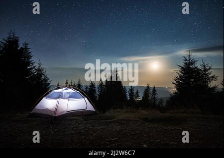 Illuminated tent setting up on edge of stone trail on mountain hill. Incredible beauty of evening landscape. Moonlight, low clouds on background of starry sky. Mountain hills in the distance. Stock Photo