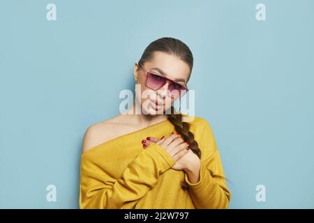 Lovely young woman presses palms to heart, feels touched and grateful, dressed in yellow jumper, isolated over blue background Stock Photo