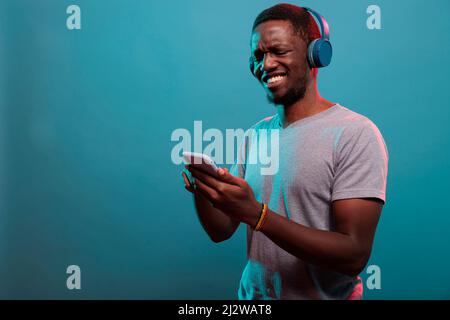 Millennial guy using smartphone and enjoying music on headphones, having fun with mp3 audio sound. Positive adult with mobile phone listening to record song on wireless headset. Stock Photo