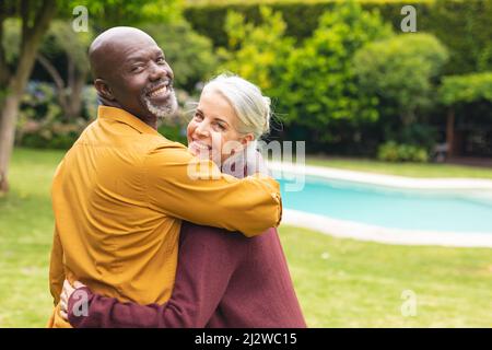 Portrait of happy multiracial senior couple looking over shoulders in backyard Stock Photo