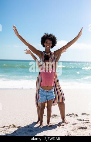 Full length portrait of happy african american mother and daughter with arms raised at beach Stock Photo