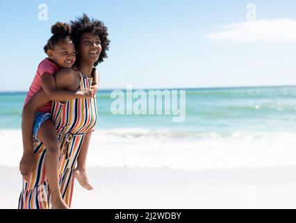 Smiling african american mother giving piggyback ride to daughter at beach on sunny day Stock Photo