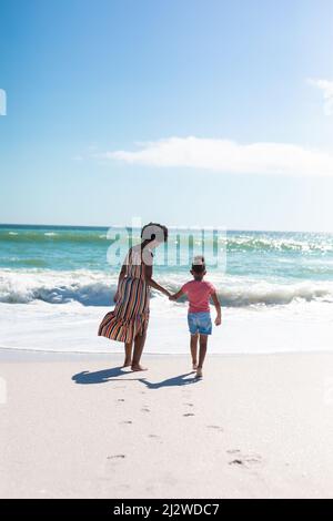 African american mother holding hand of daughter while walking towards sea at beach on sunny day Stock Photo