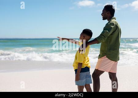 Side view of african american boy pointing while standing by father at beach on sunny day Stock Photo