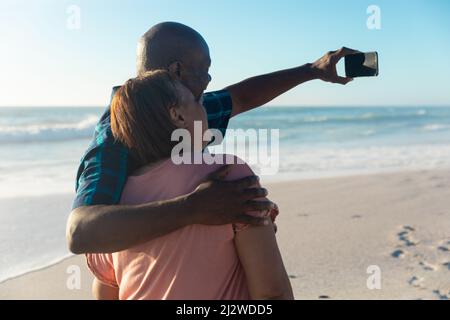 African american senior man with arm around woman taking selfie through smartphone at beach Stock Photo