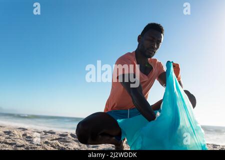 African american man crouching while collecting garbage in plastic bag at beach against blue sky Stock Photo