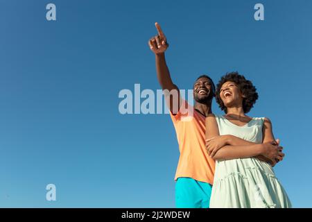 Low angle view of happy african american man pointing with girlfriend against clear blue sky Stock Photo