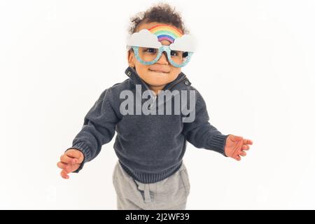 lovely curly excited toddler standing and wearing cute sunglasses with clouds and rainbow white background studio shot cowboy shot copy space. High quality photo Stock Photo