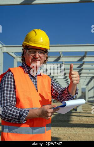 Construction Foreman Holding Clipboard and Looking at Camera. Worker in Yellow Hardhat  Standing at Construction Site and Showing Thumbs Up Hand Sign. Stock Photo