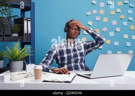 Tired and stressed customer support operator with headache in teleconversation with potential client. Fatigued consultant agent worker with burnout syndrome sitting at desk in office workspace. Stock Photo