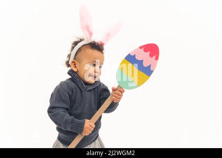 Easter and childhood concept. Happy amazed little biracial toddler boy wearing bunny ears headband and holding paper Easter egg posing over white background. High quality photo Stock Photo