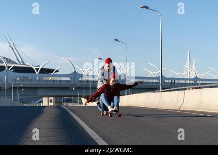 Casual couple on longboard: man teaching girl to balance on skateboard push her back down empty road Stock Photo