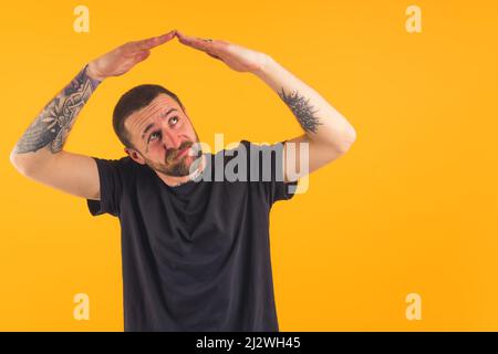 Caucasian unshaven tattooed man in a black t-shirt holding folded hands above his head like house roof looking uncertainly and mistrustfully at the imaginary roof over yellow background. High quality photo Stock Photo