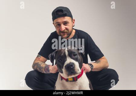 Adorable bond between pedigree dog and its owner. European bearded tattooed man in black clothes sitting in a studio with his pedigree dog. Blurred dog in the foreground. Looking at camera together. High quality photo Stock Photo