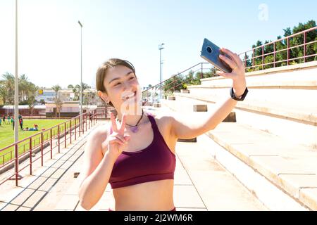 Smiling young Caucasian female athlete dressed in a purple top taking a selfie on a bleacher forming a V with her right hand. in the background is a s Stock Photo