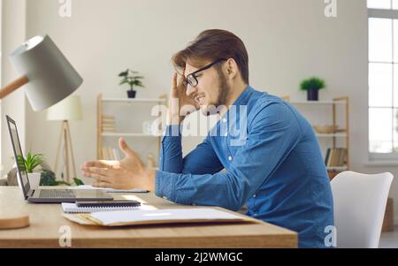 Confused and annoyed man sitting at work desk and looking at his laptop computer Stock Photo