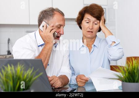 Worried mature family couple with phone sitting at laptop at kitchen table Stock Photo