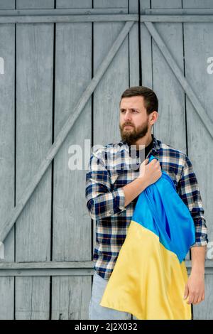 Young man holding blue-yellow State Flag of Ukraine in right hand over heart on gray wooden fence background. Guy in a checkered shirt, vertical photo Stock Photo
