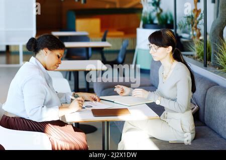 Side view portrait of two female business partners discussing project while working at table in cafe or food court Stock Photo