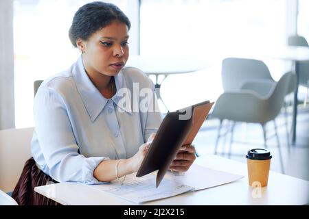 Minimal portrait of young black businesswoman using digital tablet while working at table in cafe or food court Stock Photo