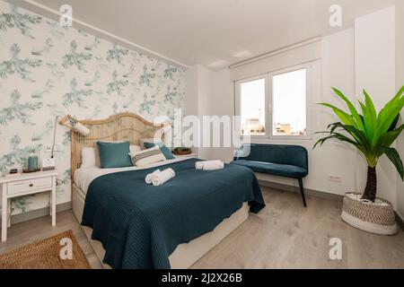 Bedroom with a double bed with a wicker headboard, walls with decorative wallpaper with plant motifs, a natural fiber rug, a blue blanket and a matchi Stock Photo