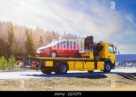 Yellow flatbed tow truck on road carrying red breakdown car on a sunny day of spring, rear and side view. Stock Photo