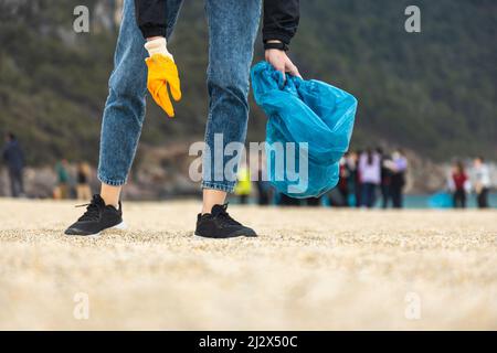 A woman in gloves with a special blue bag picks up garbage among the sand along the coast. The problem of environmental pollution. Cleaning up trash Stock Photo