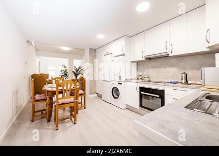 Spacious kitchen with appliances, washer and dryer stacker, gray countertops and white cabinets with light hardwood floors, dining area with pine wood Stock Photo