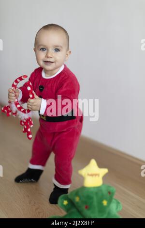 Baby girl wearing Santa's dress and deer's horns behind small Christmas tree soft toy. First Christmas Stock Photo