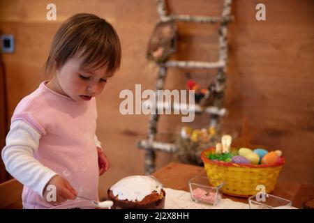 2-year-old toddler got dirty in egg white while helping decorating Easter cake. Sensory activity at home. Orthodox Easter decoration. Kids involvement Stock Photo