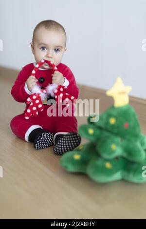 Baby girl wearing Santa's dress biting on deer's horns behind small Christmas tree soft toy. First Christmas Stock Photo