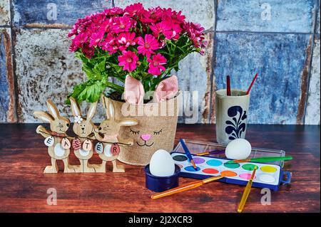 Flowerpot with a rabbit's face, wooden Easter bunnies with the text Easter on the Easter eggs and a paint box with brushes. Stock Photo