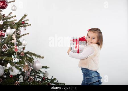 3-years old girl holding a gift box wrapped in silver paper and red ribbon. Holiday spirit photograph on white background. Christmas tree Stock Photo