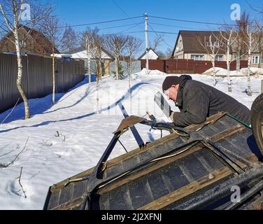 The master repairs a car trailer using electric arc welding. An elderly male worker sparks during steel welding on a spring sunny day against the backdrop of snow. Stock Photo