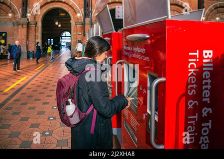 Young woman tourist buys a ticket for the metro, bus or train in a red vending machine in the main hall of the Copenhagen Central railway Station. Den Stock Photo