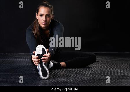 Staying supple and strong. Studio portrait of a young woman in gym clothes stretching her legs against a dark background. Stock Photo